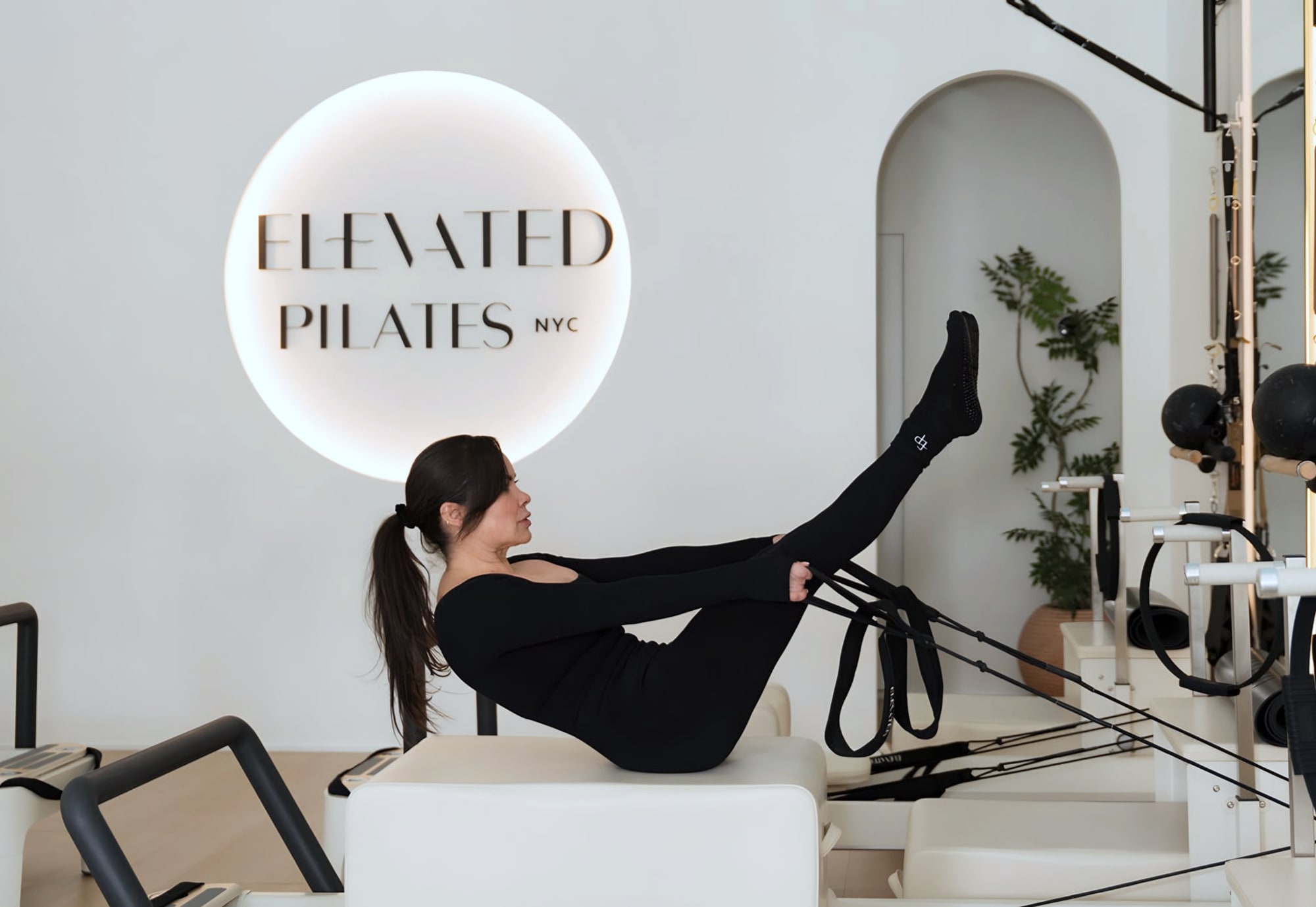 Woman on reformer holding straps beneath glowing Elevated Pilates NYC sign